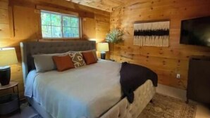 Un dormitorio tipo cabaña de madera con una cama grande, un ventilador de techo, una ventana y un televisor montado.