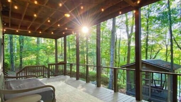 Un espacio al aire libre techado con piso de madera, una zona de estar con mesa y sillas, y vista a un área boscosa.
