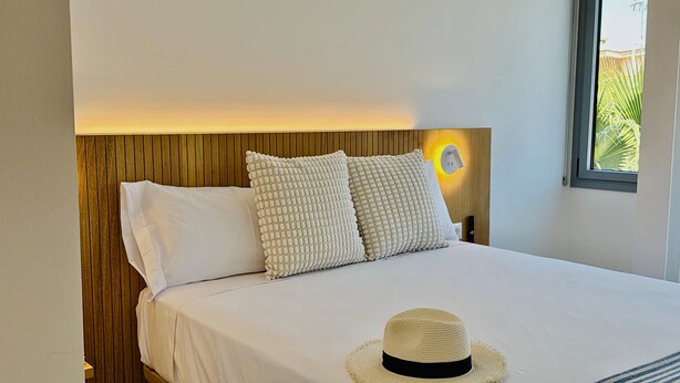 Een bed met witte beddengoed, twee gestructureerde kussens en een strohoed op het bed.
