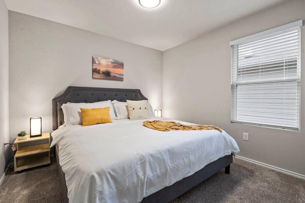 A bedroom with a large bed, a nightstand with a lamp, a window with blinds, and a painting on the wall.