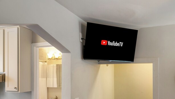 Un televisor de pantalla plana montado en una pared en una habitación con paredes blancas y una cocina visible a través de un umbral abierto.