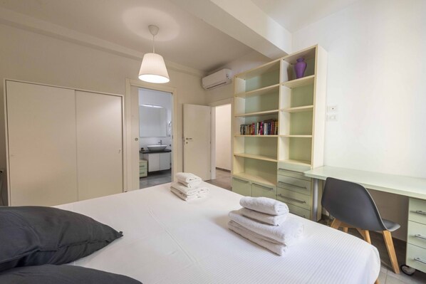 A bedroom with a bed, a desk, a chair, a bookshelf, and a bathroom visible through an open door.