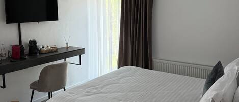 A hotel room with a bed, a desk with a flat-screen TV, a chair, a coffee maker, and a radiator.