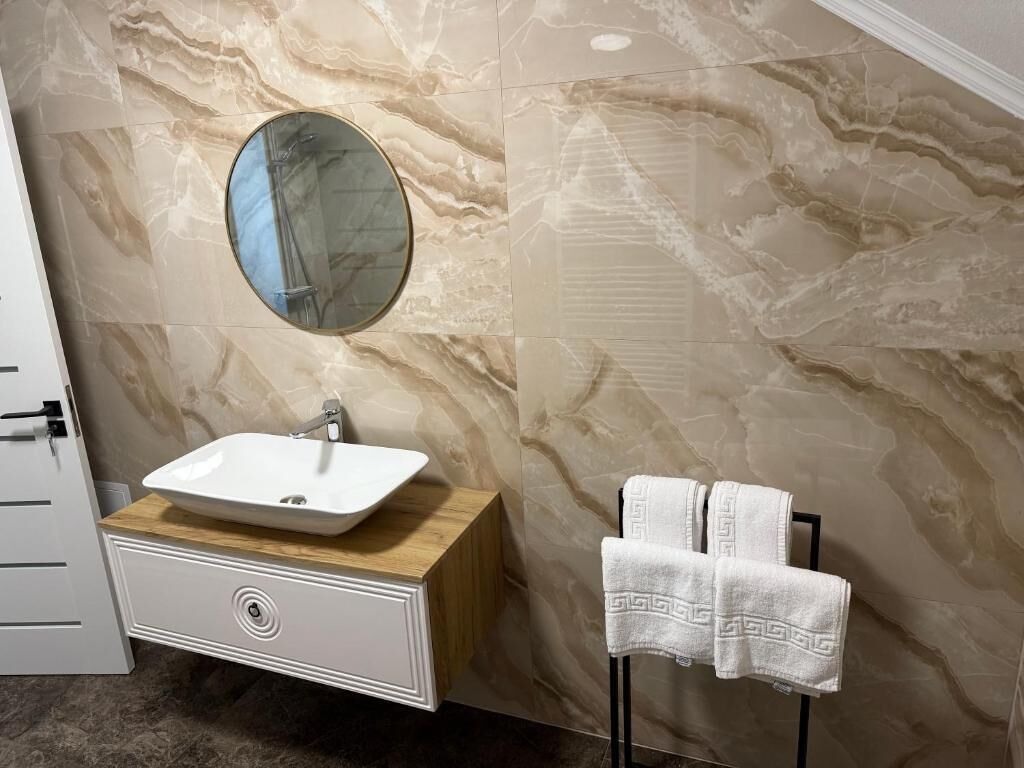 A bathroom with a marble wall, a round mirror, a white sink, and a towel rack with two towels.