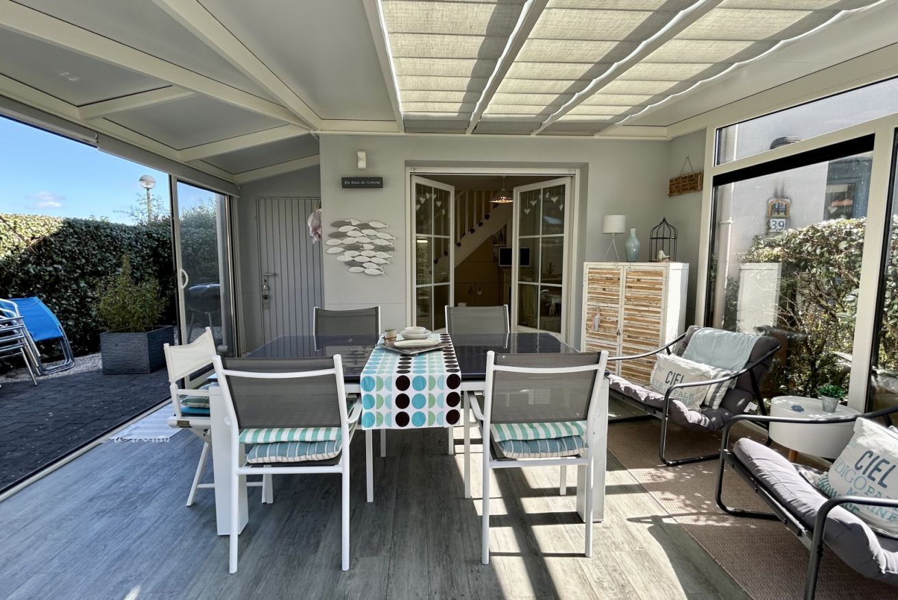 An outdoor dining area with a table and chairs, a covered seating area with cushions, and a view of the garden.