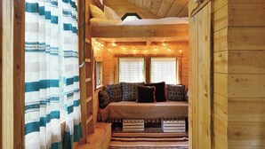 Interior acogedor de una cabaña con cama en altillo, un sofá con cojines estampados y una ventana con luces de cuerda.