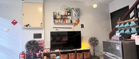 A living room with a TV, a fan, a refrigerator, and a wooden table with a remote control.
