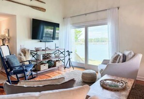 A living room with a TV, a chair, a sofa, a table with a tray, and a view of a body of water through large windows.