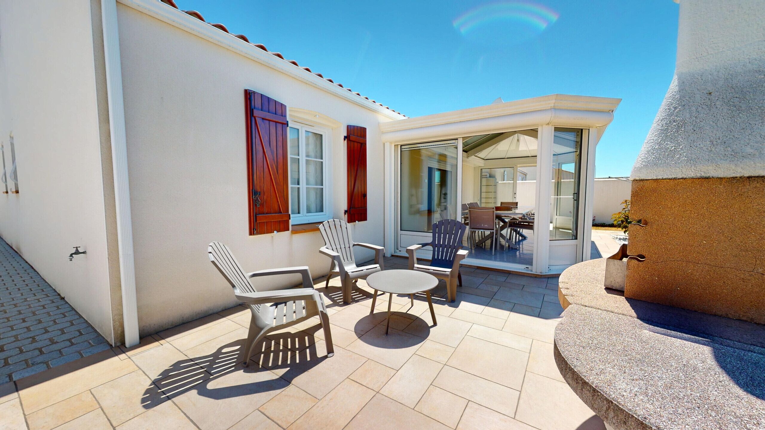 A patio with outdoor furniture, a glass door leading to an interior space, and a white building with red shutters.