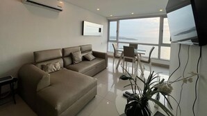 Un soggiorno moderno con un divano, una televisione a schermo piatto e vista mare attraverso ampie finestre.