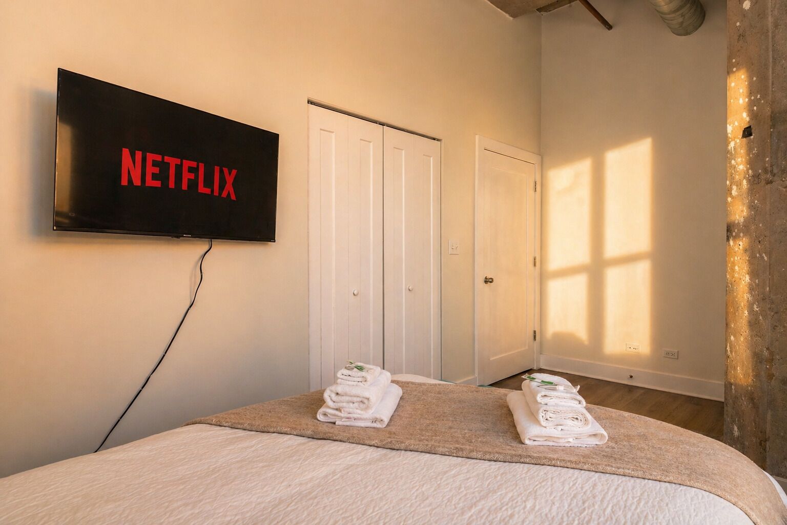 A hotel room with a bed, a TV mounted on the wall displaying 'NETFLIX,' and two doors.