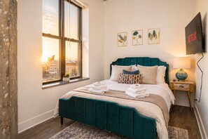 A hotel room with a large window, a bed with teal bedding, a bedside table with a lamp, and a wall-mounted TV.