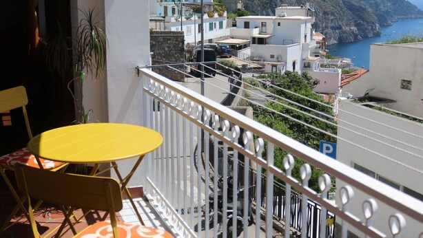 Un balcon avec des chaises jaunes et une table, donnant sur une ville côtière avec ses bâtiments et une montagne.