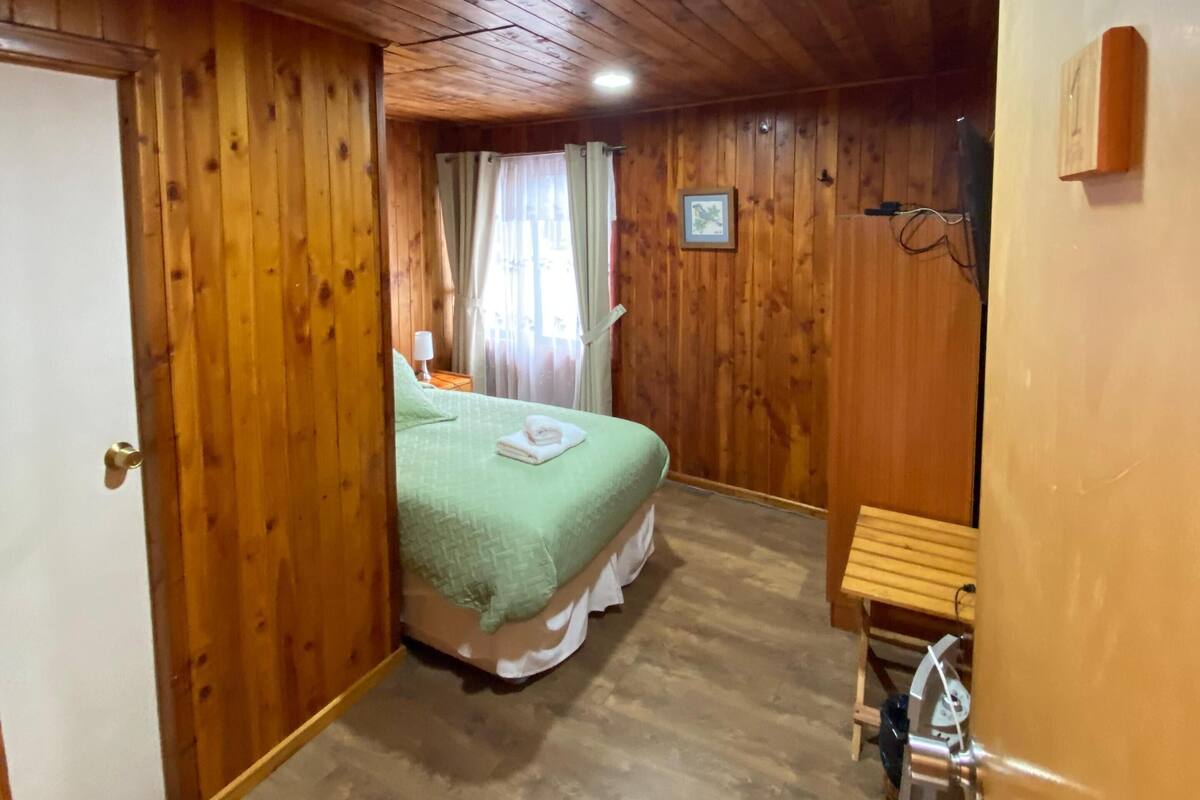 Um quarto com painéis de madeira, uma cama, uma mesinha e um banquinho.