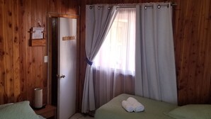 A room with wooden walls and a ceiling fan. There are two beds with green bedding and white towels. A door is visible on the left side, and a window with curtains is on the right. - Hotel Elizabeth (Panguipulli)