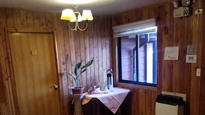 A wooden-paneled room with a door, a window, a table with a tablecloth, a potted plant, a radiator, and a rug. - Hotel Elizabeth (Panguipulli)