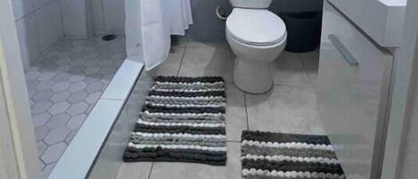 Un baño con ducha, inodoro y lavabo. Hay dos cuadros enmarcados en la pared y un perchero con toallas.