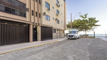 Um edifício com grandes portões de garagem, uma carrinha estacionada e um espaço pavimentado com um banco e árvores.