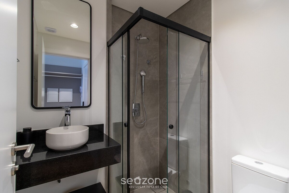 Un baño moderno con ducha acristalada, lavamanos blanco y encimera oscura.