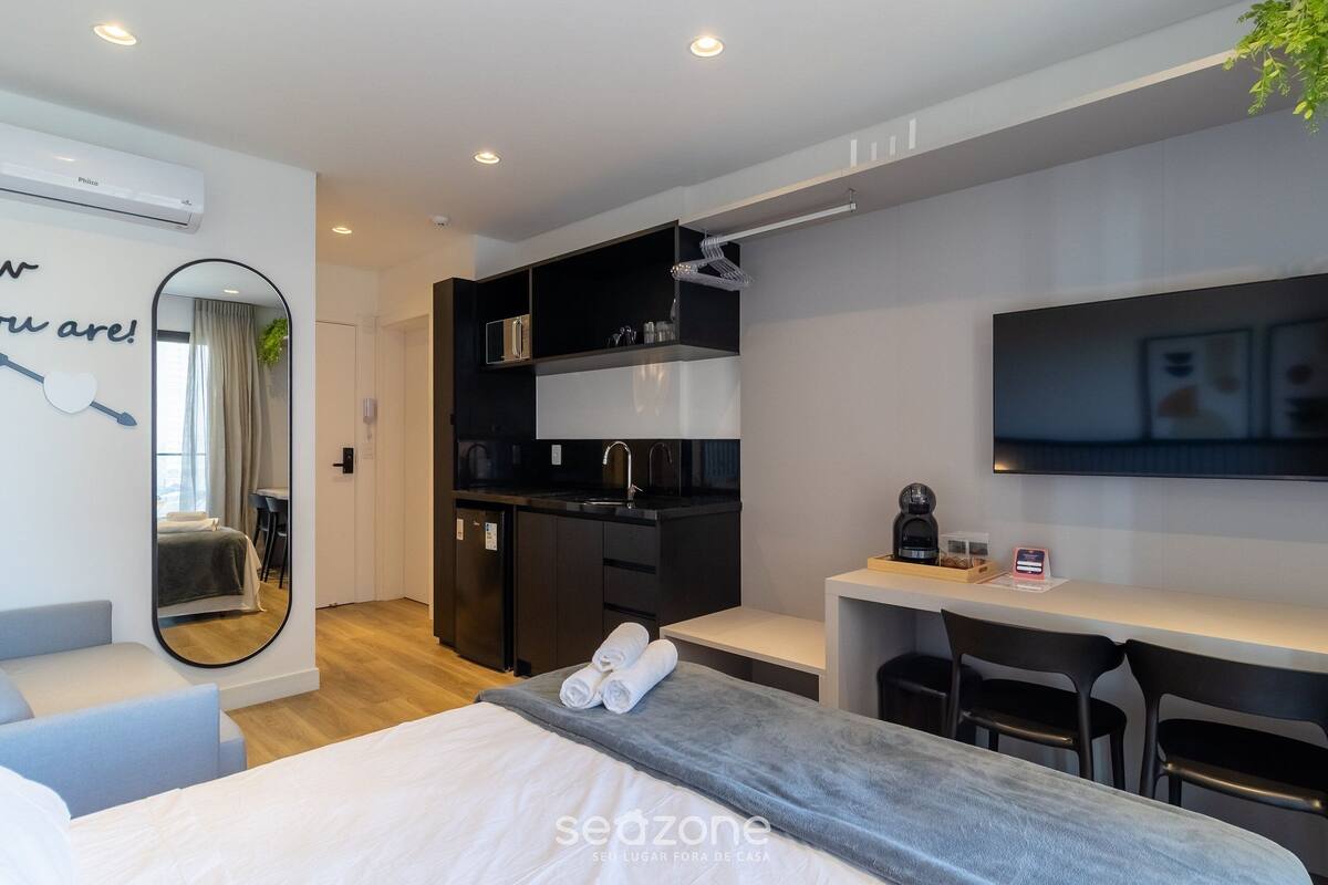 A modern hotel room with a bed, a desk, a TV, a mirror, a kitchen area, and a bathroom.