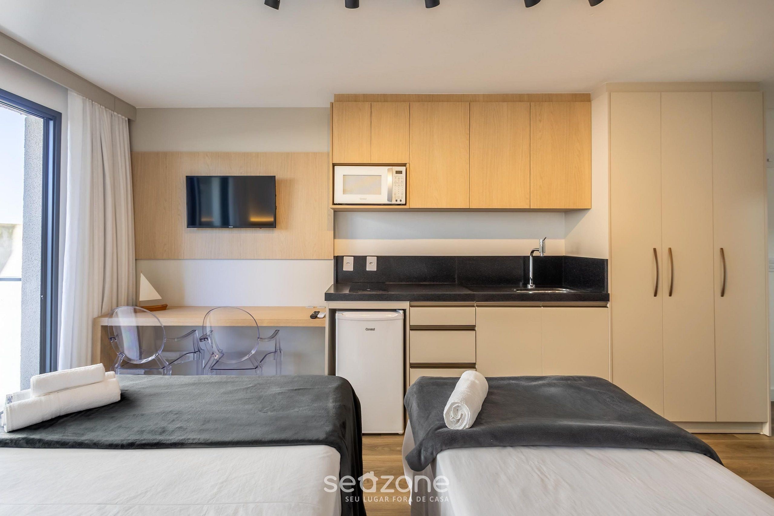 A modern hotel room with two beds, a kitchenette, a flat-screen TV, and a view of the outdoors.