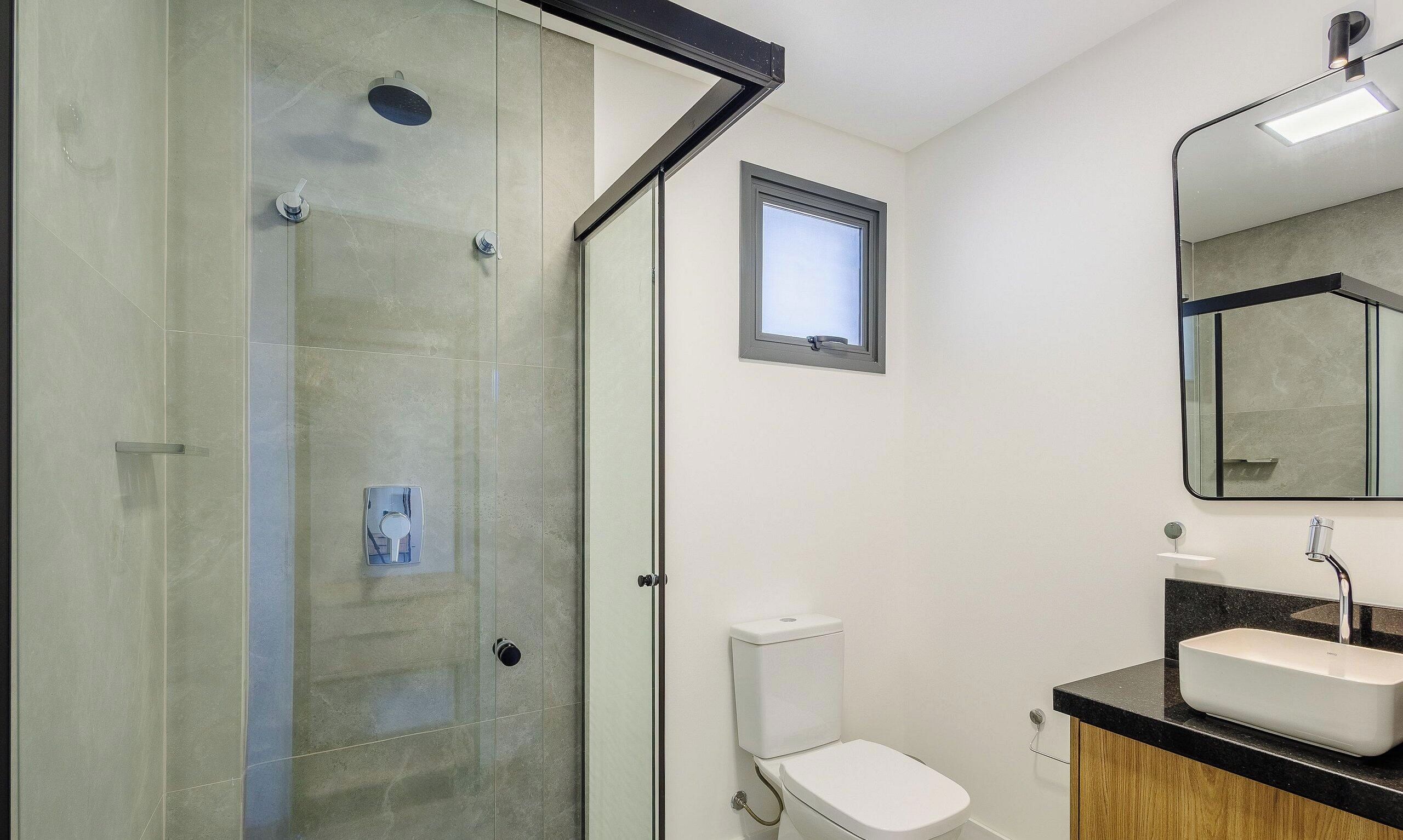 A modern bathroom with a glass shower enclosure, a toilet, a sink with a mirror, and a small window.