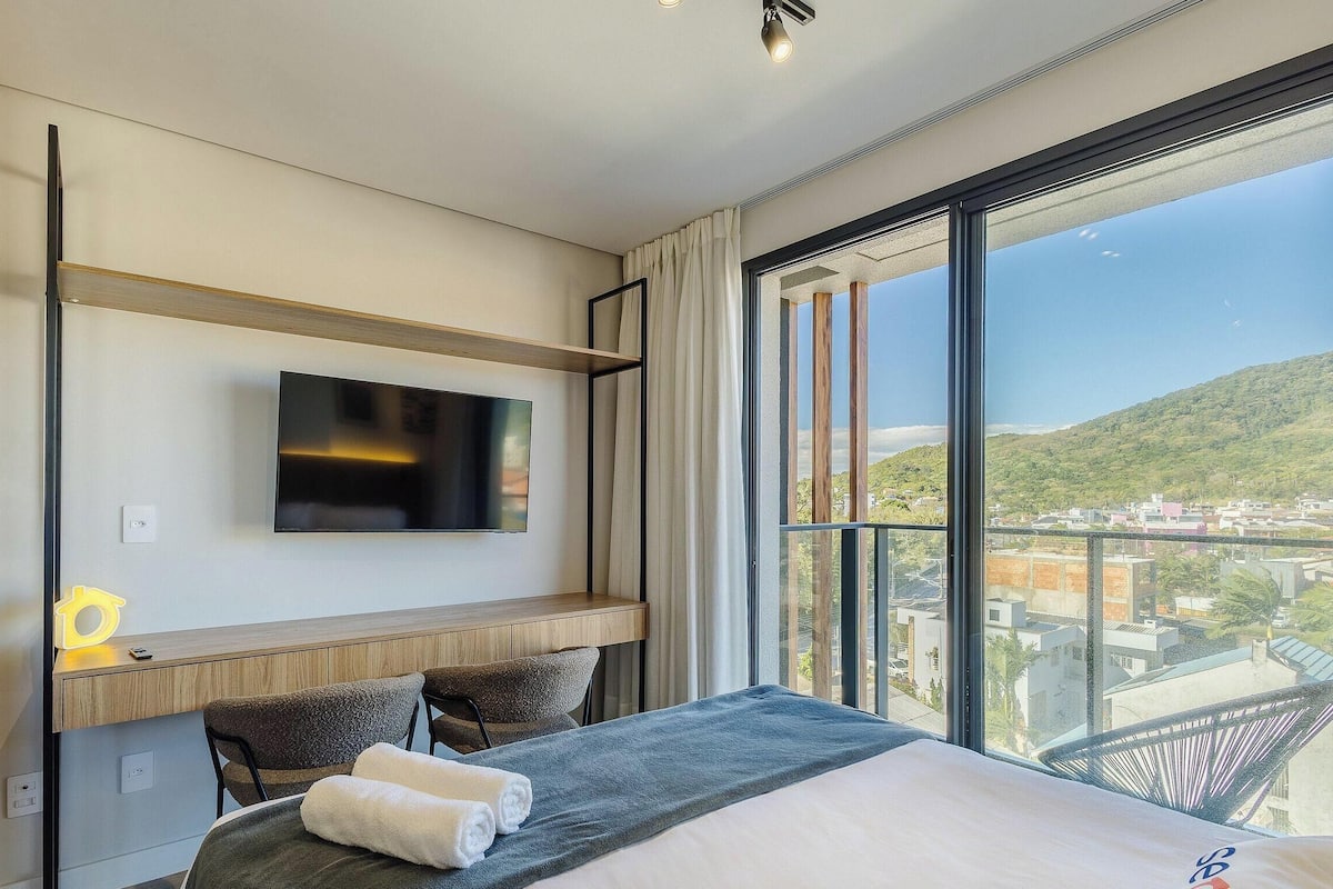 A modern hotel room with a large bed, a flat-screen TV mounted on the wall, a desk with a chair, and a balcony with a city view.