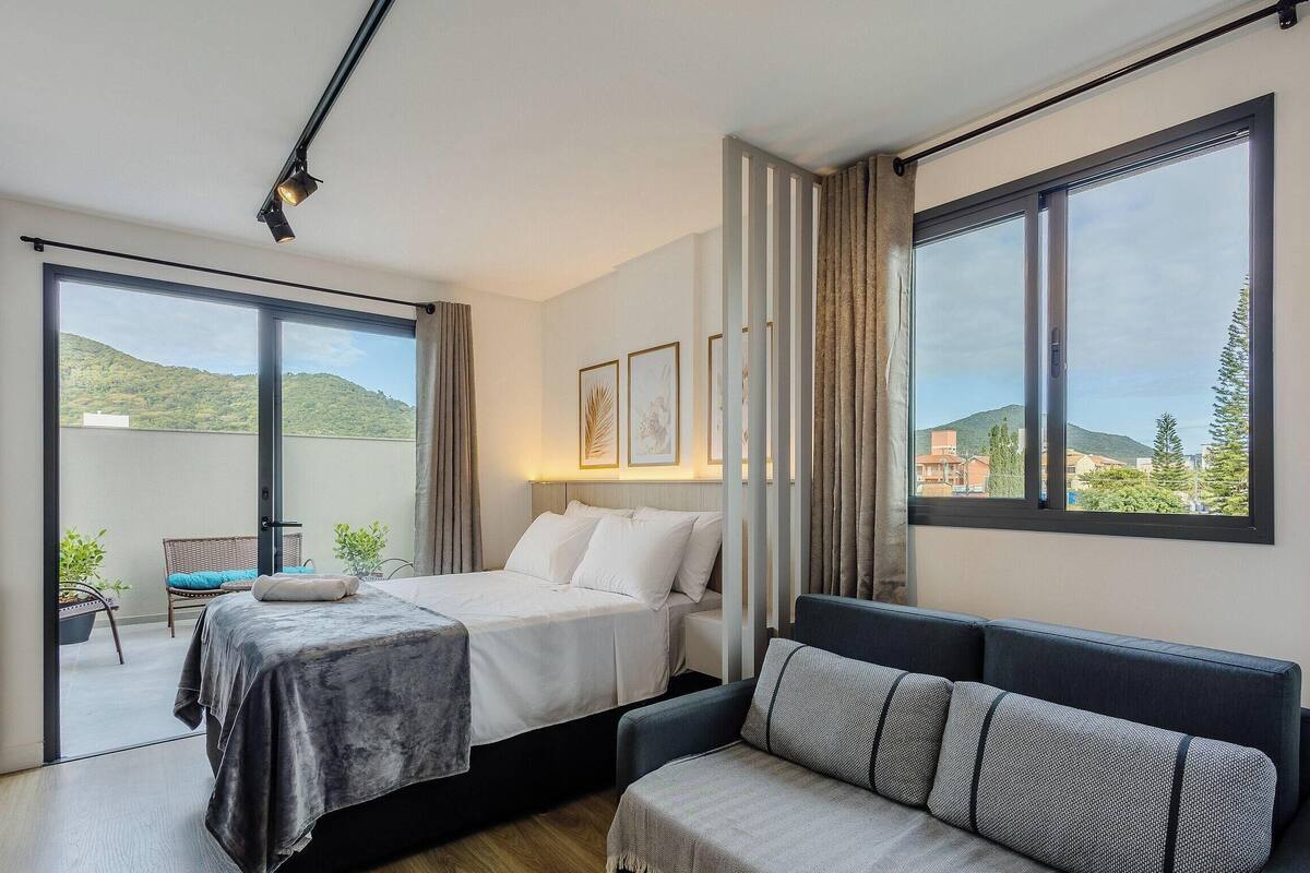 A modern bedroom with a bed, a sofa, and a balcony with a view of the hills.