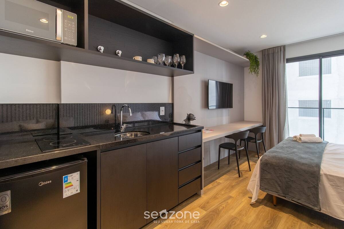A modern kitchenette with a built-in microwave, a sink, and a small refrigerator. There is a seating area with a desk and chairs, a bed with a gray blanket, and a large window providing a view.