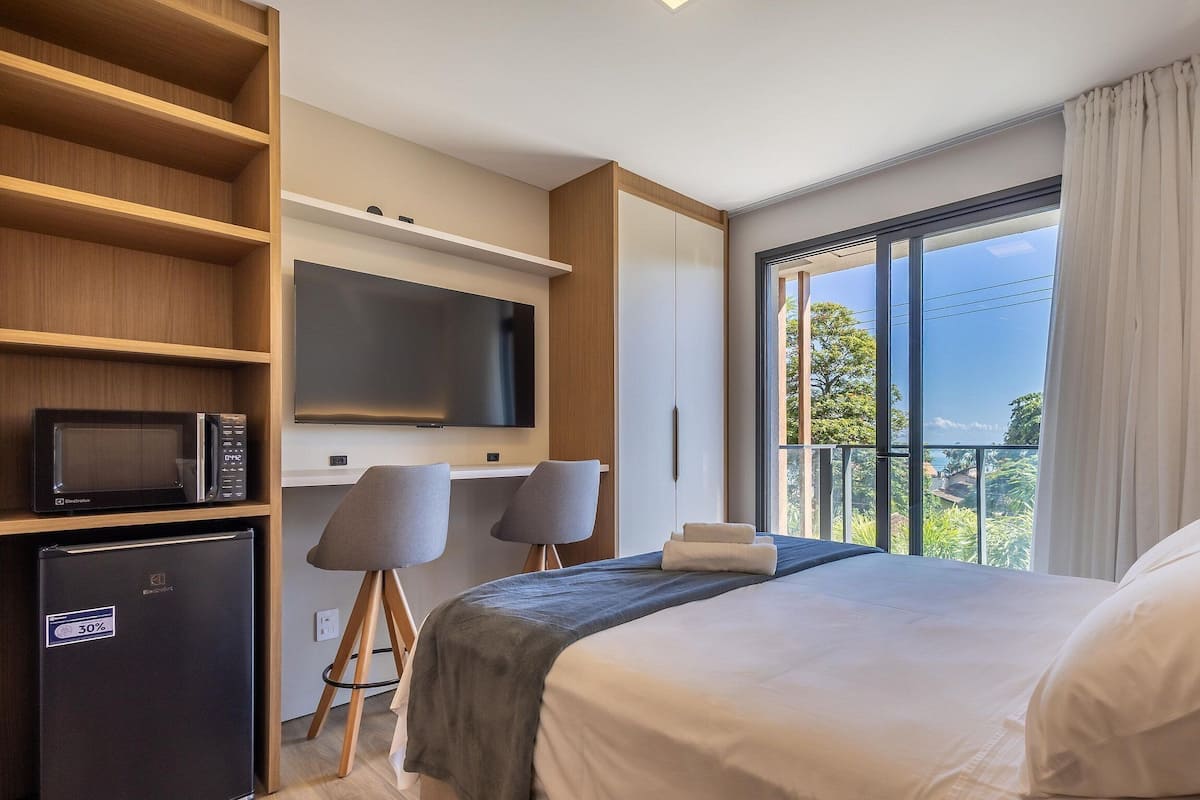A hotel room with a bed, a desk with chairs, a TV, and a balcony with a view.