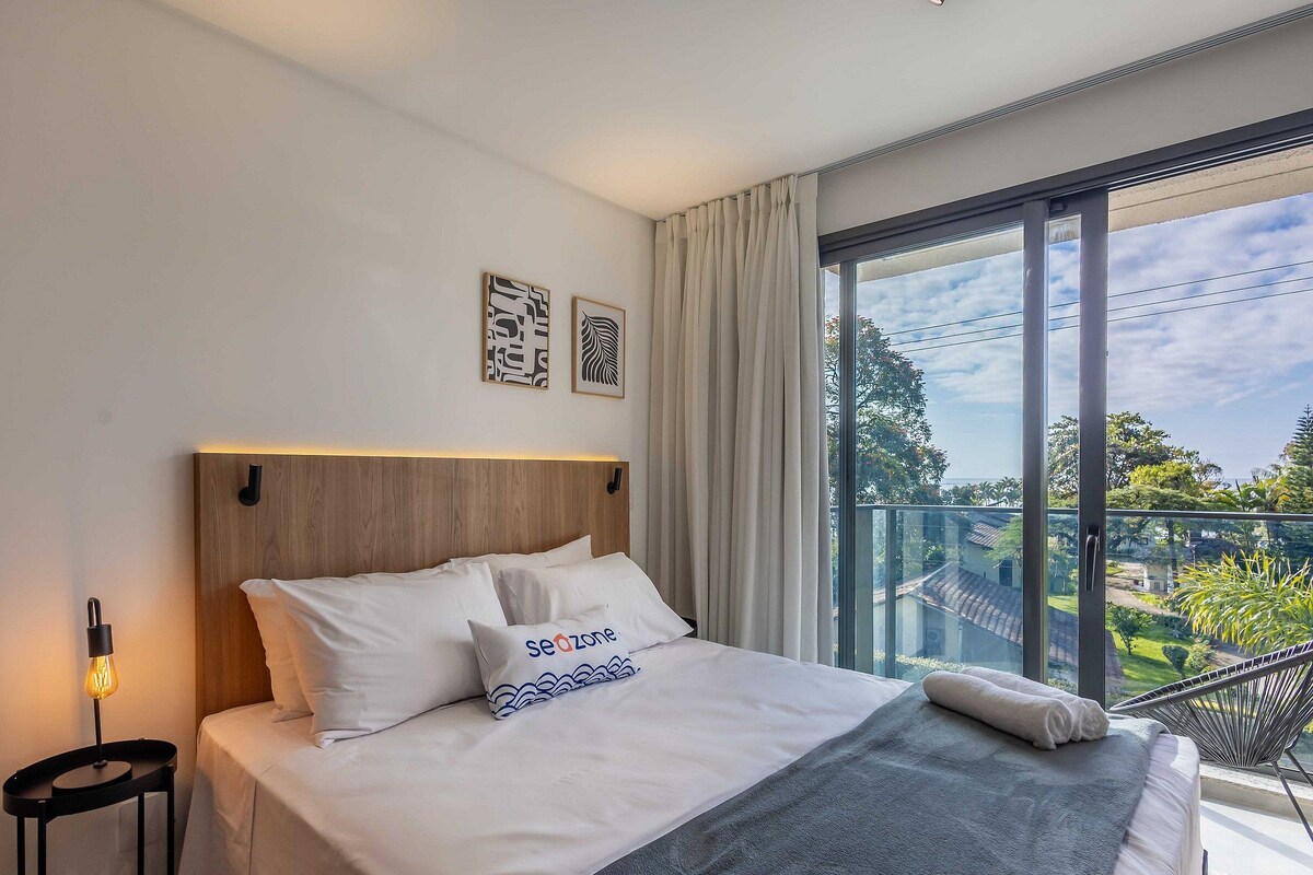 A modern hotel room with a large bed, a nightstand, a wall-mounted lamp, and a view of a balcony with greenery.