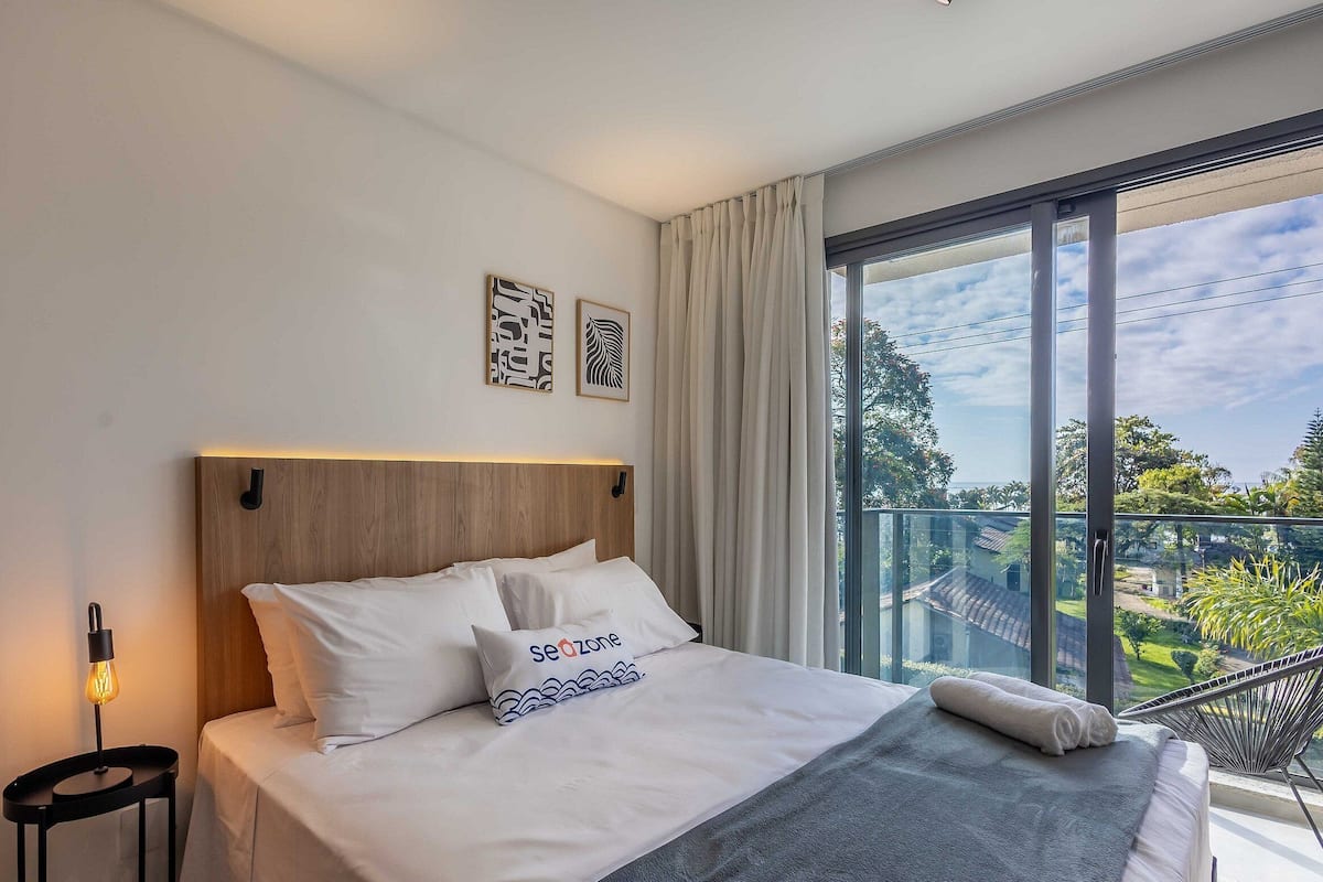 A modern hotel room with a large bed, a nightstand, a wall-mounted lamp, and a view of a balcony with greenery.