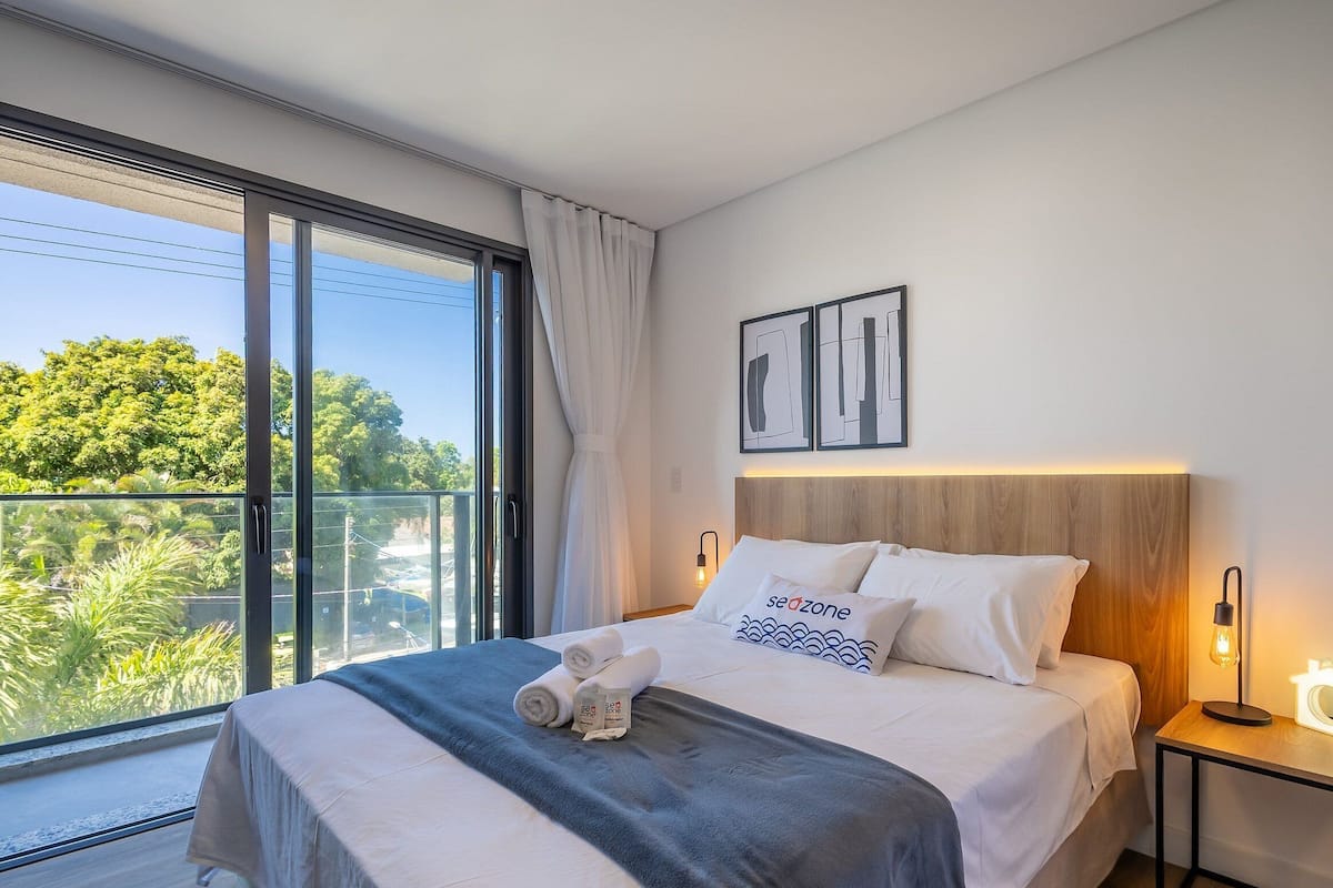 A modern hotel room with a large window, a bed with white linens, a wooden headboard, and a nightstand with a lamp.
