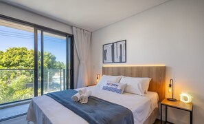 Una habitación de hotel moderna con un ventanal grande, una cama con ropa blanca, un cabecero de madera y una mesita de noche con una lámpara.