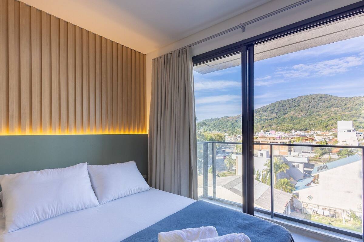 A hotel room with a large bed, a view of a cityscape, and a balcony with a mountain backdrop.