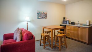 Un espacio habitable compacto con un sofá rojo, una mesa de comedor de madera con taburetes, una kitchenette con microondas y un pequeño cuadro en la pared.