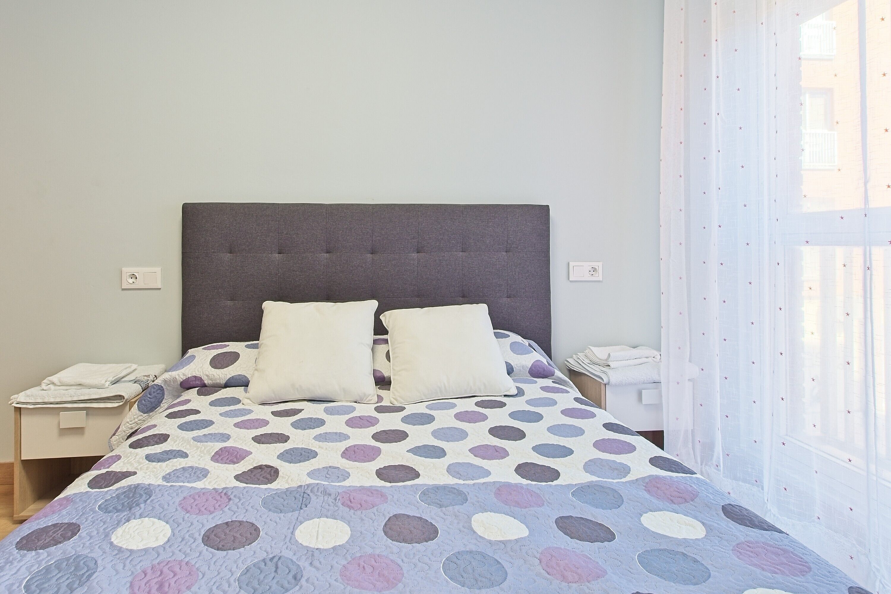 Uma cama com colcha de bolinhas, duas almofadas, cabeceira cinzenta, mesas de cabeceira com abajures e uma janela com cortinas leves.