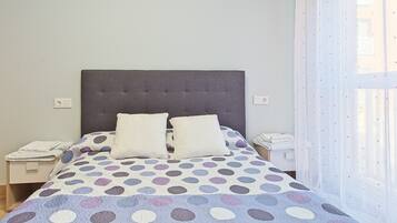 Uma cama com colcha de bolinhas, duas almofadas, cabeceira cinzenta, mesas de cabeceira com abajures e uma janela com cortinas leves.