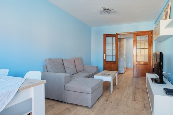 A modern living room with a grey sectional sofa, a small white coffee table, a TV on a white stand, and a wooden door leading to another room.