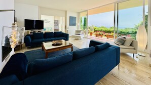 Ein modernes Wohnzimmer mit einer großen Couch, einem Couchtisch und einem Fernseher. Ein Balkon mit Blumentöpfen und Blick ins Freie ist ebenfalls vorhanden.