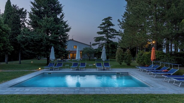 A rectangular swimming pool with lounge chairs and umbrellas, surrounded by trees and a building in the background. - La Casetta 12, Emma Villas (Sarteano)