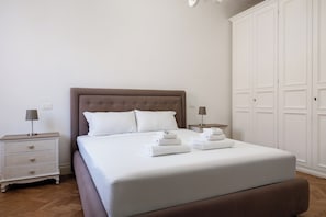 Una cama bien tendida con sábanas blancas y dos almohadas, una mesita de noche con una lámpara y un amplio armario blanco.
