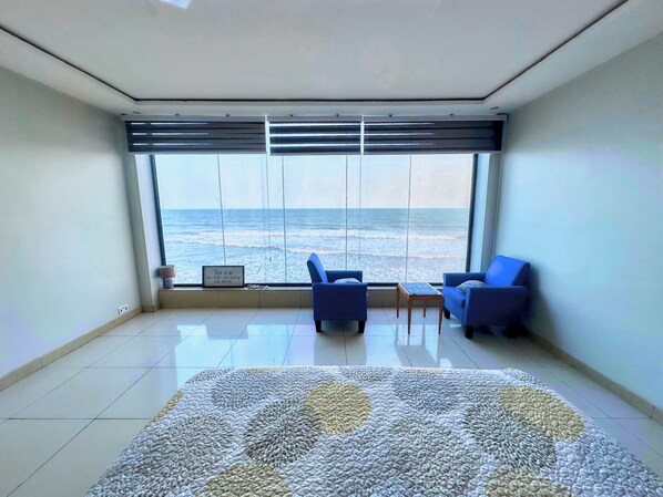 A room with a large window overlooking the ocean, two blue chairs, a small table, and a patterned rug.