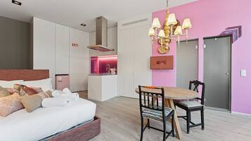 A modern hotel room with a bed, a dining table with chairs, a kitchenette, and a pink accent wall.