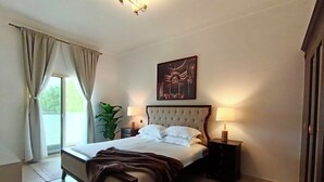 A bedroom with a large bed, two bedside tables with lamps, a window with curtains, a potted plant, and a painting on the wall.