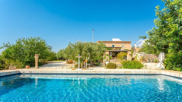 A clear blue pool with a stone border, surrounded by greenery and a building with a tiled roof. - Villa Nina Large Private Pool, A/C, WiFi (Scicli)