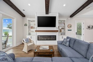 A living room with a fireplace, a large flat-screen TV, wicker furniture, and a bookshelf. - Litchfield Family Fun | Golf Cart, Spa & Huge Arcade (Pawleys Island)