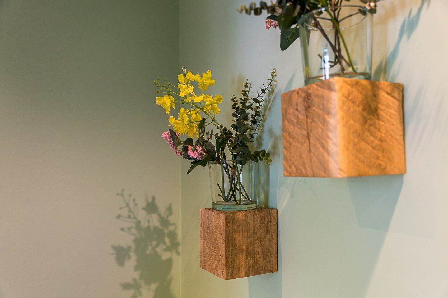 Two glass vases with flowers on a wooden block shelf against a wall.