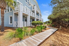 A two-story house with a wooden deck leading to the entrance, surrounded by landscaped garden beds and a palm tree.