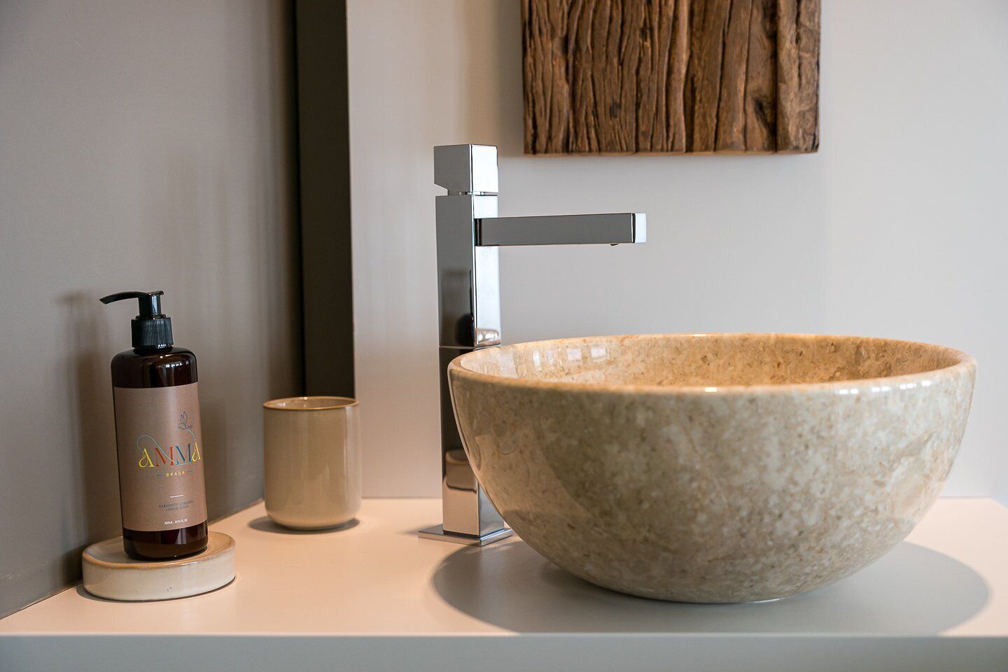 A bathroom with a large ceramic sink, a modern faucet, a bottle of shampoo, and a small cup.